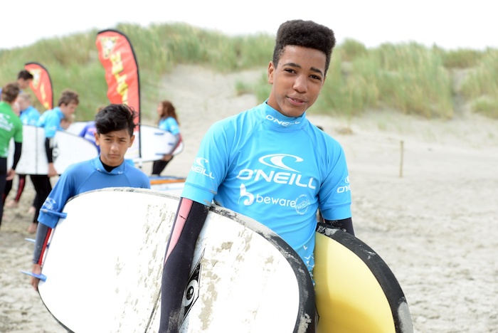 Jongen draagt surfboards en lacht naar de camera op kitesurfkamp Nederland