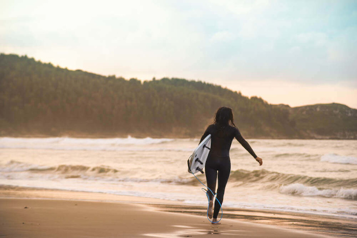Een meisje met surfboard loopt door de golven bij zonsondergang.