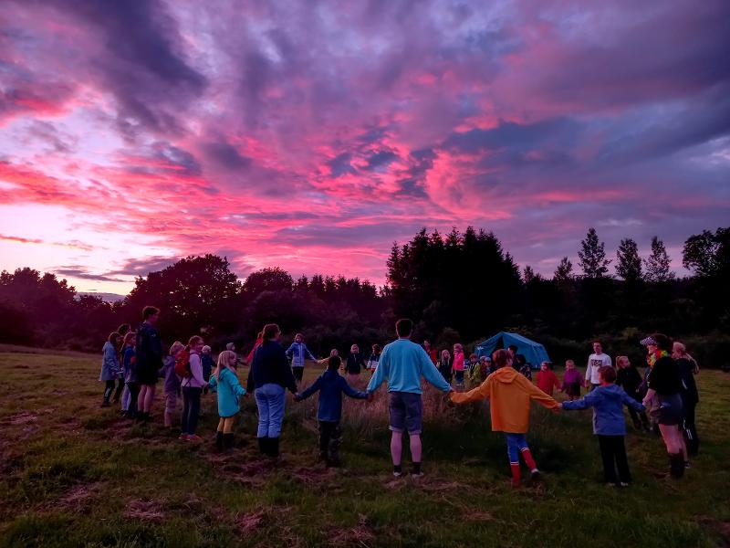 Deelnemers van vakantiekamp jeugd staan in een cirkel in een grasveld tijdens zonsondergang