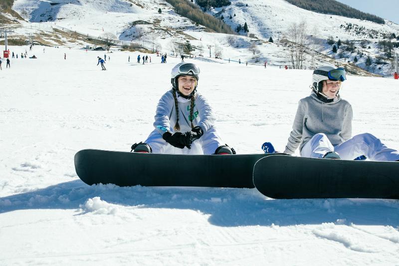 Twee meisjes met snowboards op groepsreis skivakantie zitten in de sneeuw