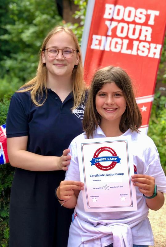 Een deelnemer van een taalvakantie Engels jeugd staat met de diploma in de handen voor een begeleider voor een vlag