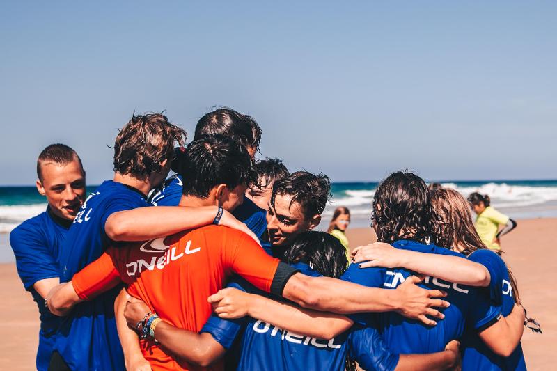 Een groep delnemers aan jongerenreis 16 jaar staan op het strand en omhelzen elkaar na het surfen