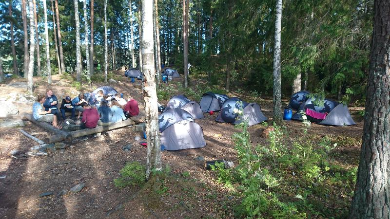een groepje deelnemers zitten bij het tentenkamp samen te eten in het bos tijdens een natuur kamp in zweden
