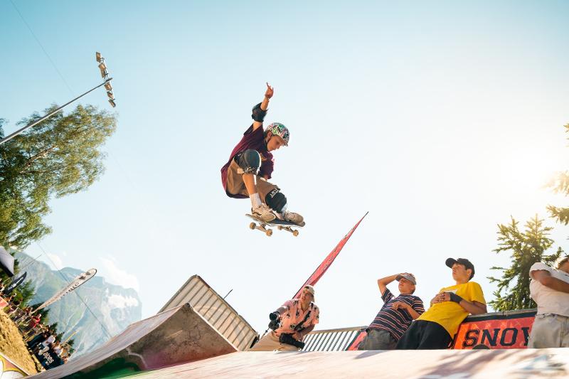  Num skate park ao ar livre na Bélgica, um jovem realiza uma manobra aérea com o skate, elevando-se acima de um corrimão. Ao fundo, vê-se uma montanha e um céu azul, criando um contraste entre a natureza selvagem e a energia urbana, tornando este campo de férias bélgica o sítio perfeito.