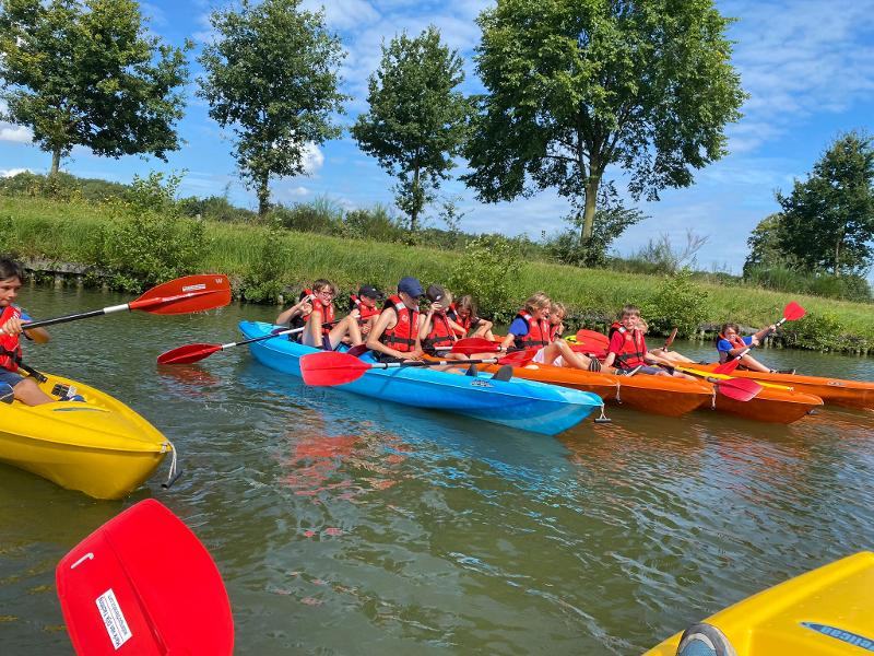  Um grupo de jovens, todos com coletes salva-vidas, desfrutam de uma atividade de caiaque num rio rodeado por árvores na Bélgica. Estão distribuídos em caiaques coloridos – amarelos, azuis e laranja – enquanto remam juntos sob um céu azul durante o campo de férias bélgica.